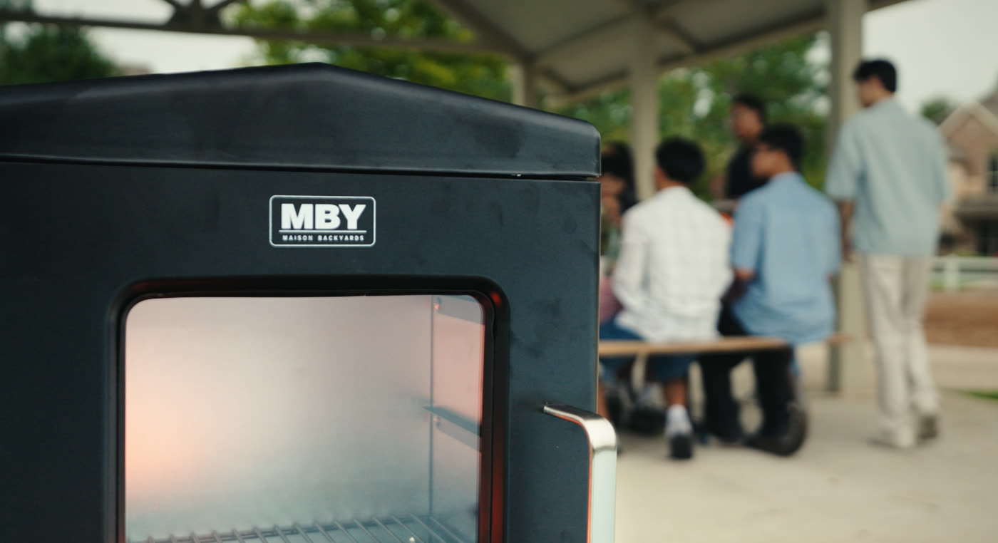 MBY-branded steel smoker on a patio with a gathering blurred in the background
